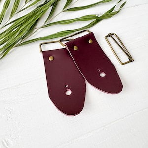 Pode incluir: Duas pontas de correia de couro bordô com fivelas de latão e rebites dourados. As pontas da correia estão sobre uma superfície branca.