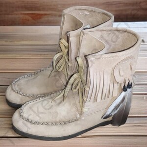 Close-up of handcrafted suede fringe moccasin boots with side feather embellishments. The boots have soft soles, beige suede leather, hand-stitched detailing, and lace-up fronts. A perfect choice for bohemian, festival, or western-themed fashion. Pictured on wood background to emphasize the natural and artisanal design of the footwear.