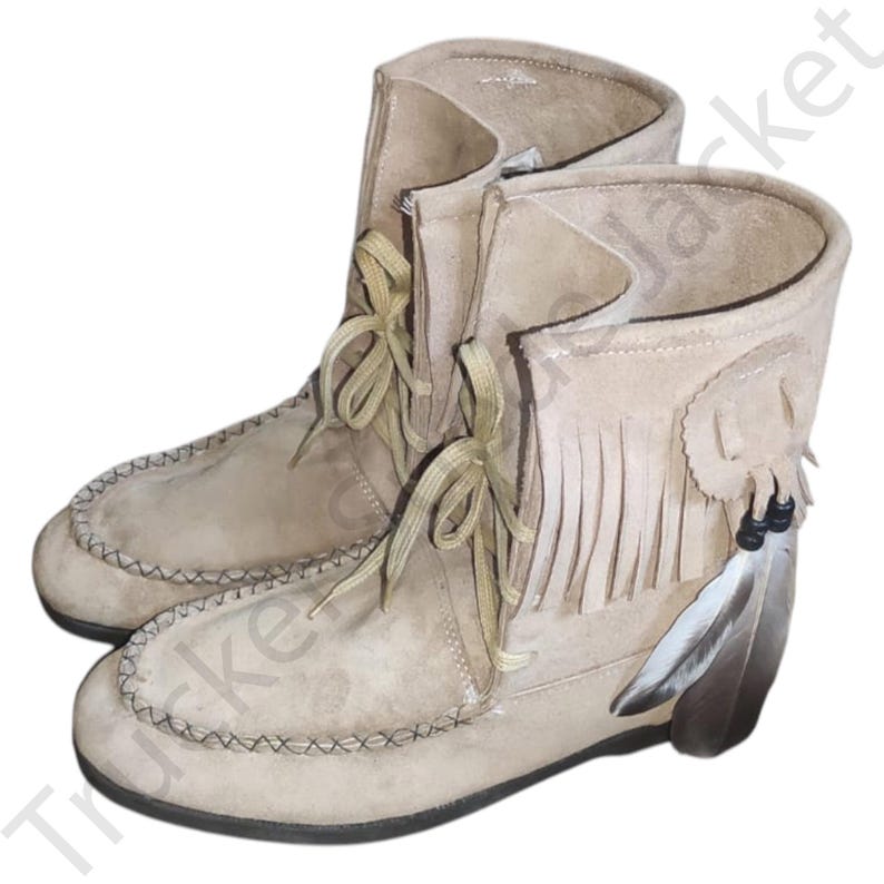 Side view of a pair of beige suede moccasin boots with fringe and decorative feathers, showcasing traditional Native-style craftsmanship. Soft sole and lace-up front make them both comfortable and stylish. Ideal for boho, rustic, or festival fashion. The image emphasizes handmade details and high-quality suede materials, presented on a natural wood background for a rustic appeal.