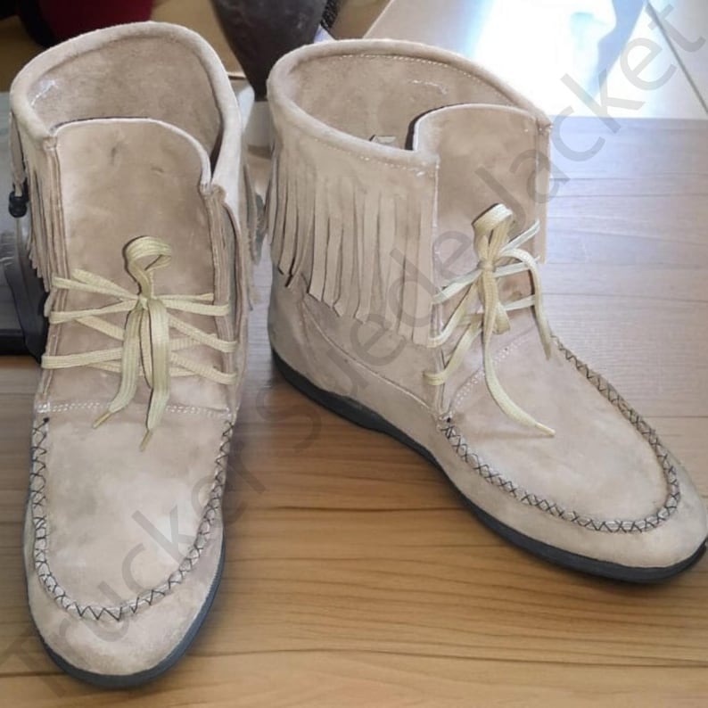 Handmade suede moccasin boots in soft beige color, featuring intricate stitching, fringe accents on the sides, and decorative natural feathers. These ankle-high boots are inspired by Native American and boho festival styles. Perfect for tribal, western, or rustic outfits. Displayed on a wooden surface to highlight the earthy tones and handcrafted texture of the footwear.