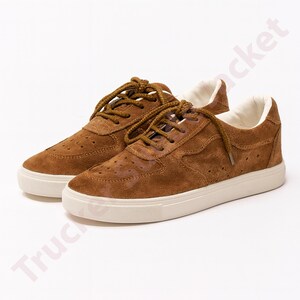 May include: A pair of brown suede trainers with white soles and brown laces. The shoes feature a perforated design on the upper and a padded white interior. The trainers are set against a white backdrop.