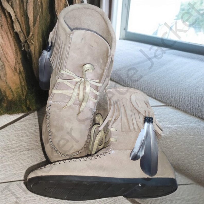 Side view of a pair of beige suede moccasin boots with fringe and decorative feathers, showcasing traditional Native-style craftsmanship. Soft sole and lace-up front make them both comfortable and stylish. Ideal for boho, rustic, or festival fashion. The image emphasizes handmade details and high-quality suede materials, presented on a natural wood background for a rustic appeal.