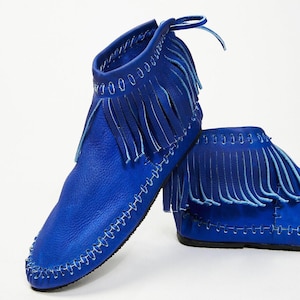 Könnte beinhalten: Ein Paar leuchtend blaue Leder-Ankle-Boots mit Fransen. Die Stiefel haben ein Mokassin-Design mit weißen Nähten an den Kanten und einer Lederschnürung oben. Der Hintergrund ist weiß, was die lebendige Farbe und die Details hervorhebt.