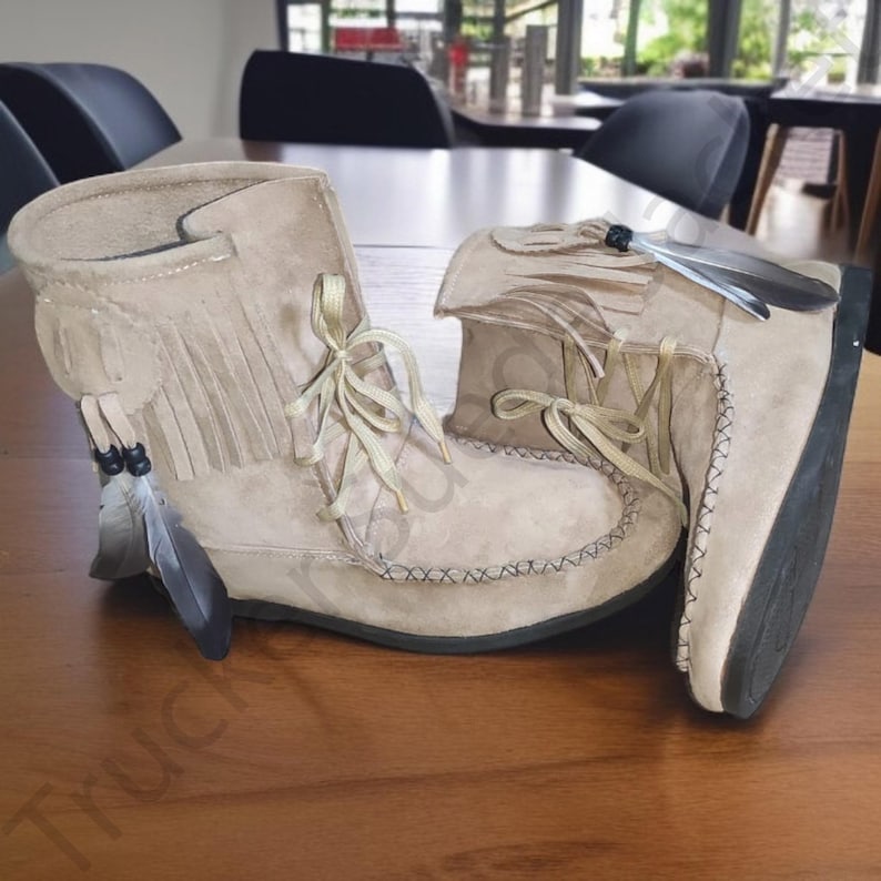 Side view of a pair of beige suede moccasin boots with fringe and decorative feathers, showcasing traditional Native-style craftsmanship. Soft sole and lace-up front make them both comfortable and stylish. Ideal for boho, rustic, or festival fashion. The image emphasizes handmade details and high-quality suede materials, presented on a natural wood background for a rustic appeal.