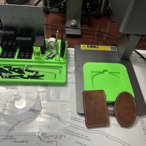 May include: A green and black tool organizer with a variety of tools and screws. There are also three brown leather patches on a gray surface.