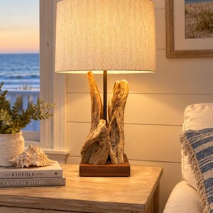May include: A table lamp featuring a cylindrical lampshade with a beige herringbone pattern, supported by a base made of natural driftwood pieces arranged vertically on a square dark brown wooden stand, emitting a warm, soft glow.