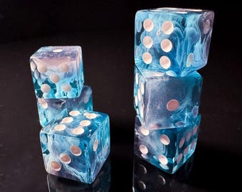 May include: Six blue and white marbled dice with white dots are stacked on a reflective black surface. The dice are translucent, allowing the marbled pattern to be visible. The dice are a gaming accessory.