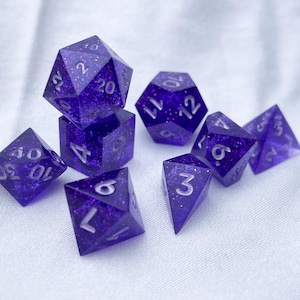 May include: A set of eight purple dice with white numbers. The dice are made of a clear resin with glitter embedded in it. The dice are in various shapes, including a d4, d6, d8, d10, d12, and d20.