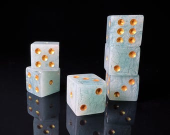 May include: Several translucent blue-green dice with gold-colored dots are stacked and arranged on a reflective black surface. The dice are cube-shaped, with each face displaying a different number of dots.