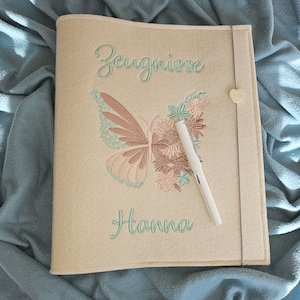 Könnte beinhalten: Cremefarbener Filzordner mit gesticktem Schmetterling und floralem Design. Der Ordner hat das Wort "Zeugnisse" und den Namen "Hanna" in Türkis gestickt. Ein weißer Stift liegt auf dem Einband, und ein herzförmiger Verschluss sichert den Ordner.
