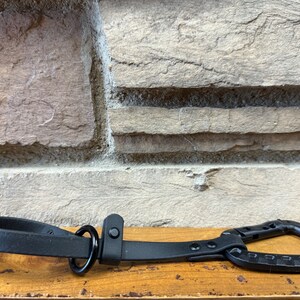 May include: Black leather dog leash with a metal carabiner clip and a metal ring. The leash is on a wooden surface.