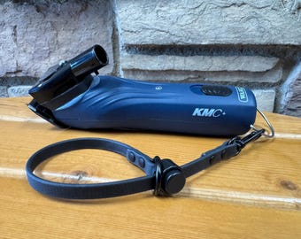 May include: A blue Wahl KMC+ hair clipper with a black cutting head and a black leather strap. The clipper is on a wooden surface. The clipper has the text "KMC+" printed on it.