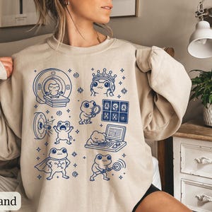 May include: A sand-colored crewneck sweatshirt featuring a dark blue graphic print of various frogs in medical and scientific scenarios, including a frog in an MRI machine, a frog wearing a crown, and a frog examining medical imaging scans.