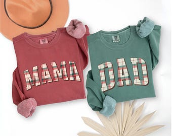 May include: Two long-sleeved sweatshirts, one red and one green, with the words "MAMA" and "DAD" in a plaid design. A tan hat and scrunchies are also visible. The sweatshirts are likely part of a family matching set.