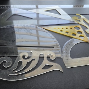 May include: A collection of clear plastic drafting tools, including triangles, protractors, and French curves, arranged on a dark surface. Some tools have measurement markings in inches and angles.