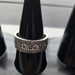 Può includere: Un anello in metallo color argento caratterizzato da un motivo in rilievo continuo di linee vorticose e piccoli punti lungo la fascia centrale. L'anello ha un profilo piatto e una finitura testurizzata e ossidata che mette in risalto i dettagli.