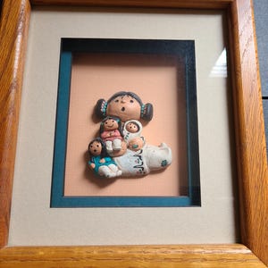 Puede incluir: Escultura en relieve enmarcada que representa a una madre y sus hijos. Las figuras son de color claro, con detalles en negro, rosa y azul. El marco es de madera y tiene un color marrón claro. El fondo es de color melocotón claro.