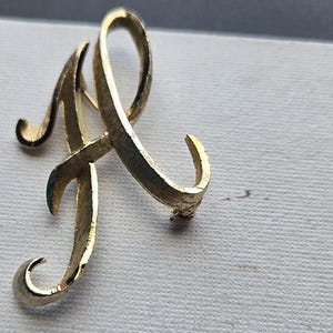 May include: A gold-coloured metal brooch in the shape of the letter "A" in a cursive script. The brooch has a curved design with a textured surface and is set against a white background.