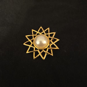 May include: A gold-toned brooch in the shape of a starburst, featuring a large, round, off-white pearl at its center. The brooch has a geometric design with pointed edges. The background is a solid black.
