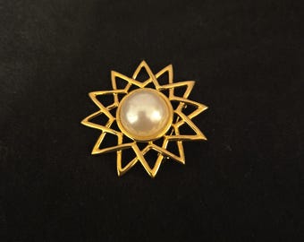 May include: A gold-toned brooch in the shape of a starburst, featuring a large, round, off-white pearl at its center. The brooch has a geometric design with pointed edges. The background is a solid black.