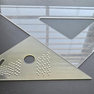 Puede incluir: Dos reglas triangulares de plástico transparente. Una regla tiene un recorte triangular, la otra un agujero y marcas. Las reglas están sobre una superficie oscura.