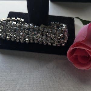 May include: A silver-toned bracelet with multiple rows of sparkling rhinestones is displayed on a black velvet stand. A pink rose with a green stem and leaves is positioned to the right of the bracelet. The bracelet is a jewelry item.