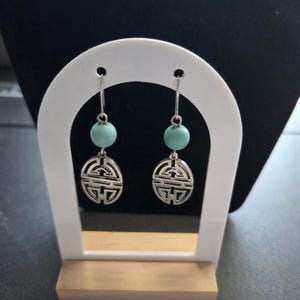 Peut inclure: Paire de boucles d'oreilles pendantes argentées. Chaque boucle d'oreille est ornée d'une perle ronde bleu clair et d'un pendentif ovale ajouré avec un motif géométrique. Les boucles sont présentées sur un présentoir blanc.
