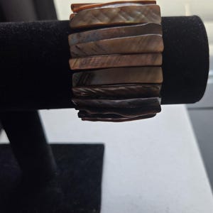 Puede incluir: Una pulsera ancha y marrón hecha de piezas rectangulares e iridiscentes de concha. La pulsera se exhibe en un expositor de joyas de terciopelo negro, mostrando su diseño en capas y sus variaciones de color naturales. Las piezas de concha tienen un aspecto pulido y liso.