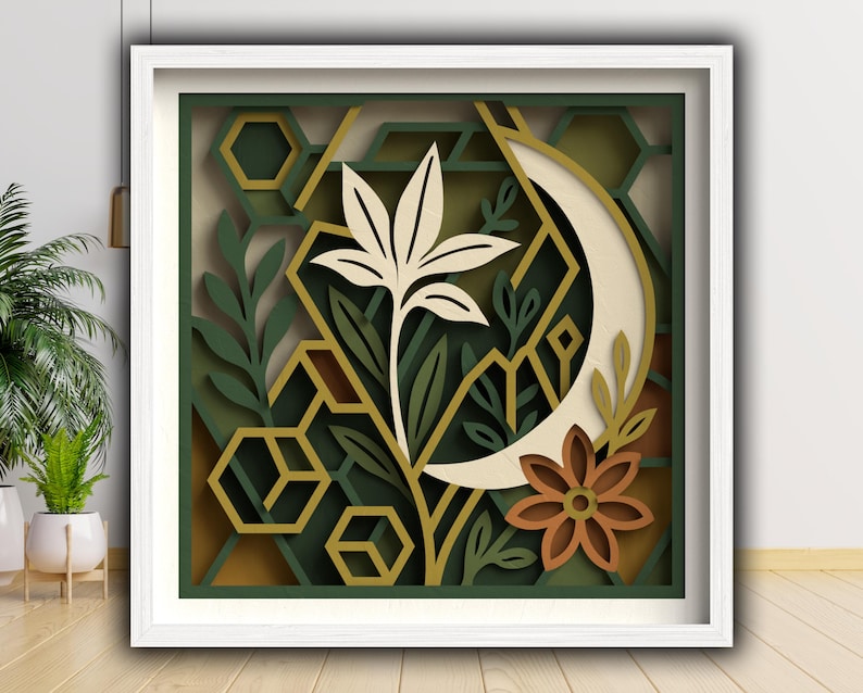 May include: A framed paper art piece with a layered botanical design. It features a cream-coloured flower, a crescent moon, and geometric shapes in green, gold, and brown. The artwork is presented within a white frame.