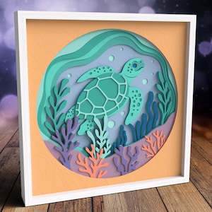 May include: A framed paper art piece featuring a sea turtle swimming in an ocean scene. The artwork uses layers of paper in shades of teal, blue, and coral to create a 3D effect. The background is a light orange color.