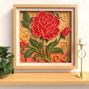 Op de afbeelding: Een ingelijst papierkunstwerk met een grote rode roos met groene bladeren en een knop. Het kunstwerk staat tegen een gele en perzik achtergrond met decoratieve krullen, alles in een licht houten frame. Een gouden kandelaar met een witte kaars staat rechts.