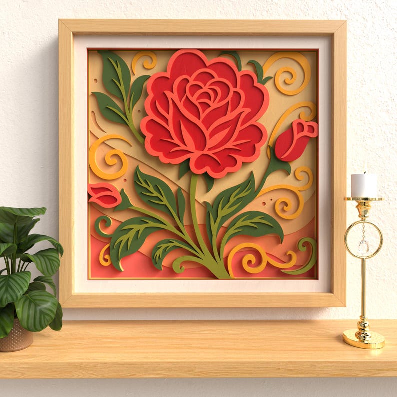 Op de afbeelding: Een ingelijst papierkunstwerk met een grote rode roos met groene bladeren en een knop. Het kunstwerk staat tegen een gele en perzik achtergrond met decoratieve krullen, alles in een licht houten frame. Een gouden kandelaar met een witte kaars staat rechts.