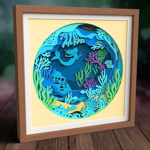 May include: A square framed wall art piece featuring a circular layered paper cutout design of an underwater scene. The artwork displays a mermaid silhouette, coral reefs, and seaweed in shades of blue, purple, and green against a light yellow background.