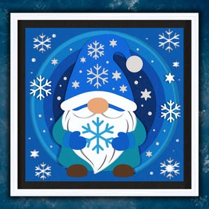 May include: A framed paper art piece featuring a winter gnome design. The gnome wears a blue hat with snowflakes and holds a snowflake. The background is layered with shades of blue and white, with additional snowflakes.