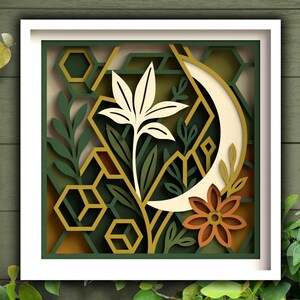 May include: A framed paper art piece with a layered design. It features a cream-coloured flower, a crescent moon, and geometric shapes in green, gold, and brown. The artwork is set against a green wooden background.