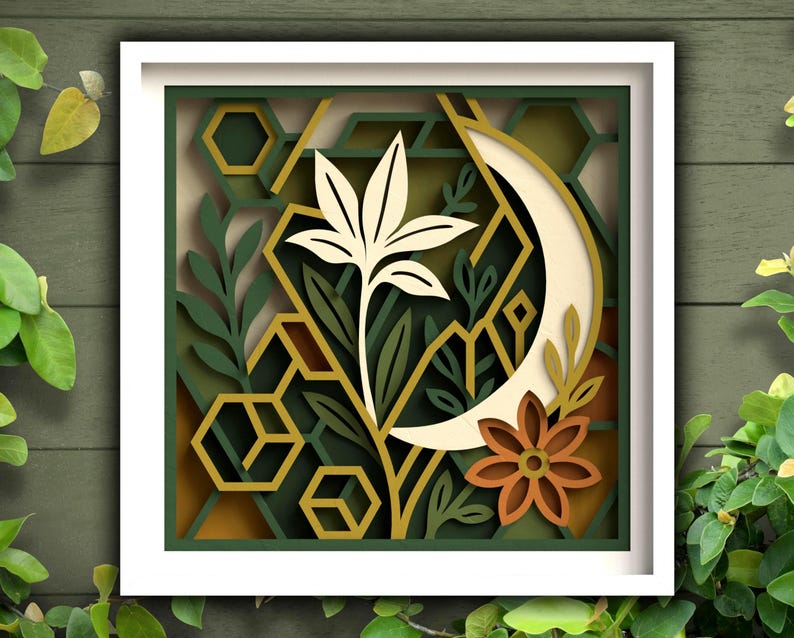 May include: A framed paper art piece with a layered design. It features a cream-coloured flower, a crescent moon, and geometric shapes in green, gold, and brown. The artwork is set against a green wooden background.