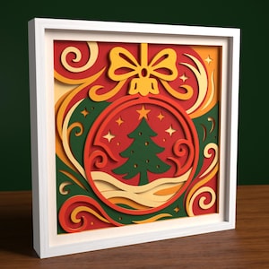 Op de afbeelding: Een ingelijst, gelaagd papieren kunstwerk met een kerstbalontwerp. De bal is rood met een groene kerstboom en sterren binnenin. De achtergrond heeft krullen en een gele strik. Het frame is wit.