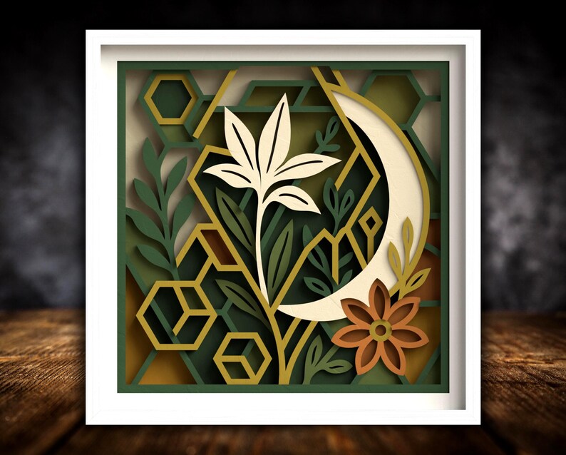 May include: A framed paper artwork with a layered botanical design. It features a cream-coloured flower, a crescent moon, and geometric shapes in green, gold, and brown. The artwork is set within a white frame.