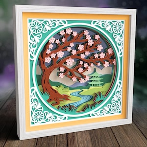 May include: A square, white-framed layered paper art piece featuring a circular cutout. The scene depicts a brown cherry blossom tree with pink flowers, a winding blue river, green hills, and a pagoda structure, all set against a yellow background.