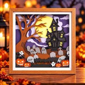 Può includere: Un'opera d'arte in carta stratificata con cornice quadrata in legno che raffigura una scena di Halloween con una casa stregata nera, una grande luna gialla, un albero marrone, lapidi grigie con la scritta RIP, zucche arancioni e fantasmi.