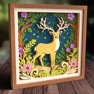 May include: A framed, layered paper art piece depicting a yellow deer with large antlers, set against a dark blue background with stars. The artwork includes green, purple, and pink floral and foliage elements, all within a wooden frame.