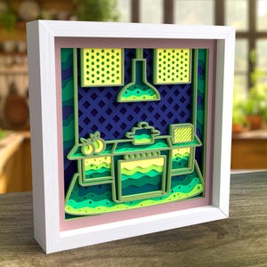 Puede incluir: Un marco blanco con un paspartú rosa que rodea una obra de arte de papel en capas que representa una escena de cocina. La escena presenta una estufa con una olla, una lámpara colgante y un fondo con estampado verde y amarillo.