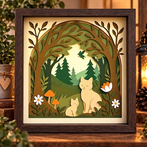 May include: A square shadow box frame containing a multi-layered paper cut art piece. The scene features a light-colored cat and kitten sitting in a forest with brown trees, green hills, pine trees, orange mushrooms, and white flowers.