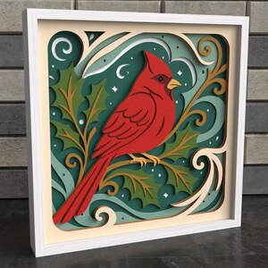 May include: A layered wooden art piece featuring a vibrant red cardinal perched on a branch with green holly leaves. The artwork is set against a teal background with white accents and is framed in a white wooden frame. The design has a 3D effect.