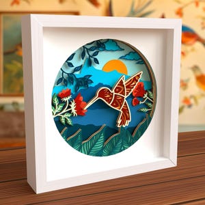 May include: A white framed wall art piece with a circular cutout featuring a papercraft hummingbird in flight, surrounded by flowers and foliage. The background is a blue and green gradient with a sun peeking out from behind the mountains.