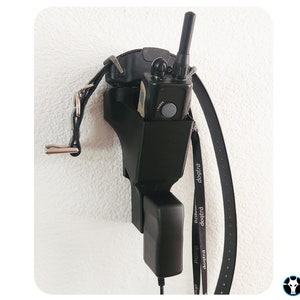 May include: Black wall mount with a black dog training collar and a black dog training device with a black antenna. The device is plugged into a black power adapter. The wall mount has a hook for hanging a leash.