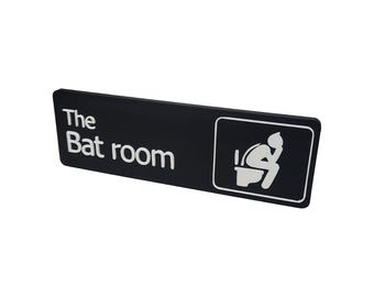 May include: Black rectangular sign with white text that reads "The Bat room" and a white silhouette of a person sitting on a toilet.