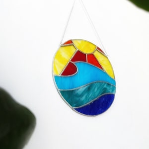 Peut inclure: Vitrail représentant un soleil et un océan. Le design ovale présente des couleurs jaune, rouge et bleu, suspendu par une chaîne argentée. Le soleil est jaune avec des détails rouges, et l'océan a des vagues bleues et turquoise.