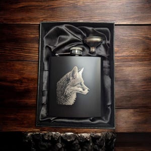 May include: A black hip flask featuring a detailed fox head engraving, presented in a black gift box with a satin lining. Includes a small funnel. The flask is approximately 10 cm tall.