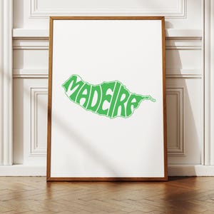 Puede incluir: Un cuadro enmarcado con la palabra "MADEIRA" en verde, con la forma del contorno de la isla. La obra de arte está sobre un fondo blanco dentro de un marco de madera, expuesta sobre un suelo de madera.